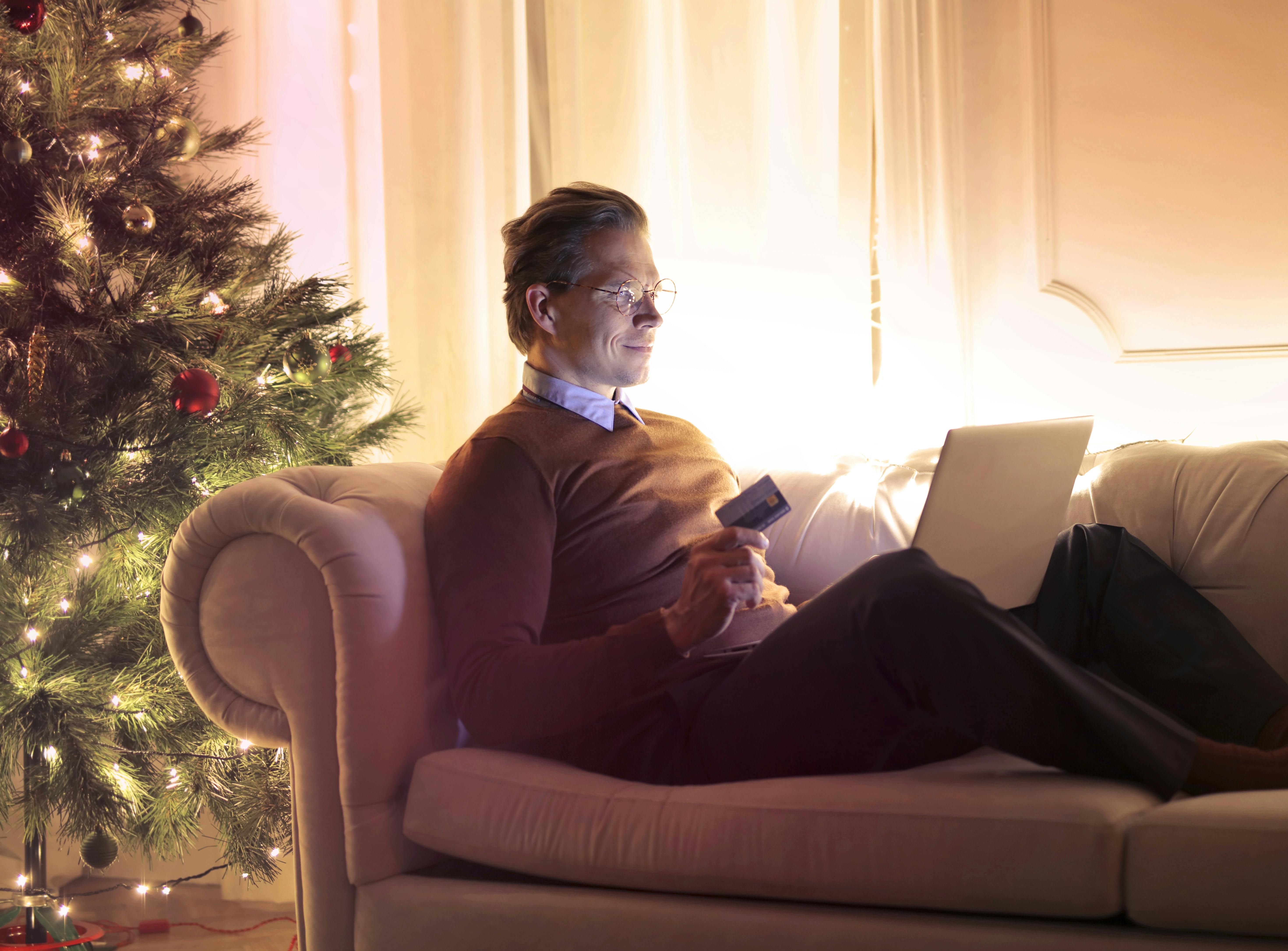Man lounging on sofa chair using laptop with Christmas tree behind him, online holiday shopping concept
